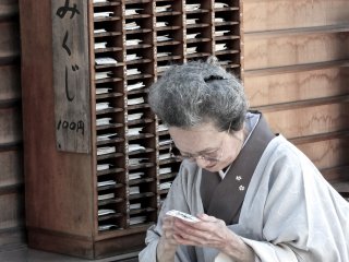 Người phụ nữ trong bộ kimono đang đọc quẻ xăm omikuji mà bà đã mua từ giá gỗ phía sau