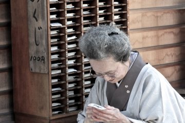 <p>Woman in kimono reading the omikuji fortune that she purchased from the wooden rack behind her</p>
