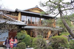 Sebuah bangunan Jepang dua lantai dengan pintu kaca, yang membuat Anda dapat melihat dan mengagumi keindahan taman Jepang. Seorang wanita dengan kimono sedang mengantri untuk masuk ke dalamnya.