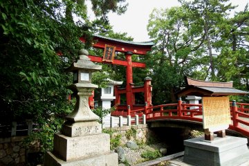 <p>Main entrance to the shrine with red torii gate, pretty bridge, and stone lanterns surrounded by green trees</p>