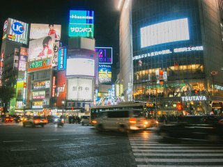 Terlihat papan reklame besar memenuhi area Shibuya Crossing, terlihat ramai di malam hari.