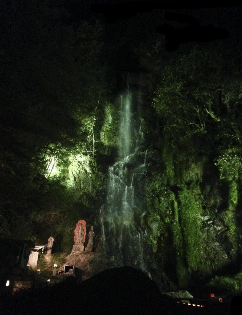 <p>An orange statue of&nbsp;Fudō-Myō-ō (immovable&nbsp;wisdom king) standing steadfast in front of the illuminated green waterfall.</p>