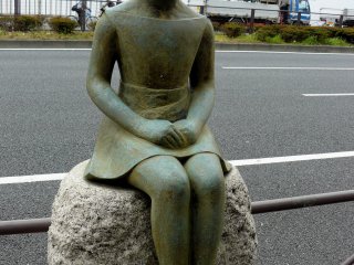 Бронзовая девочка сидит на камне возле дороги