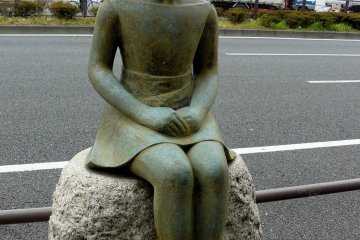 <p>Бронзовая девочка сидит на камне возле дороги</p>