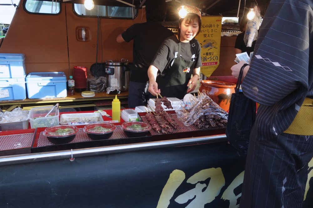 A man in traditional yukata festival wear buys two meat skewers from the "yakiniku" lady