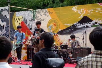 야외 무대에서, 대학의 로컬 락 밴드는 예비 학생들과 학부모들 앞에서 몇 곡을 연주해 선보인다.