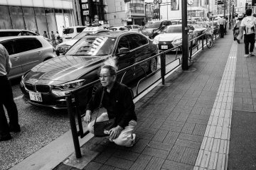 A Japanese middle-aged man squats down by the sidewalk and take a smoke break.