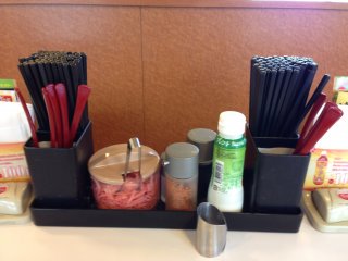 Chopsticks and spoons are readily available at the tables and counters in Sukiya, as are condiments, the ticket holder, and the ordering bell